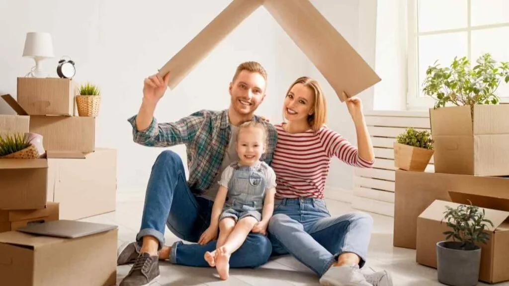 Casal e criança segurando um telhado de papelão entre caixas de mudança